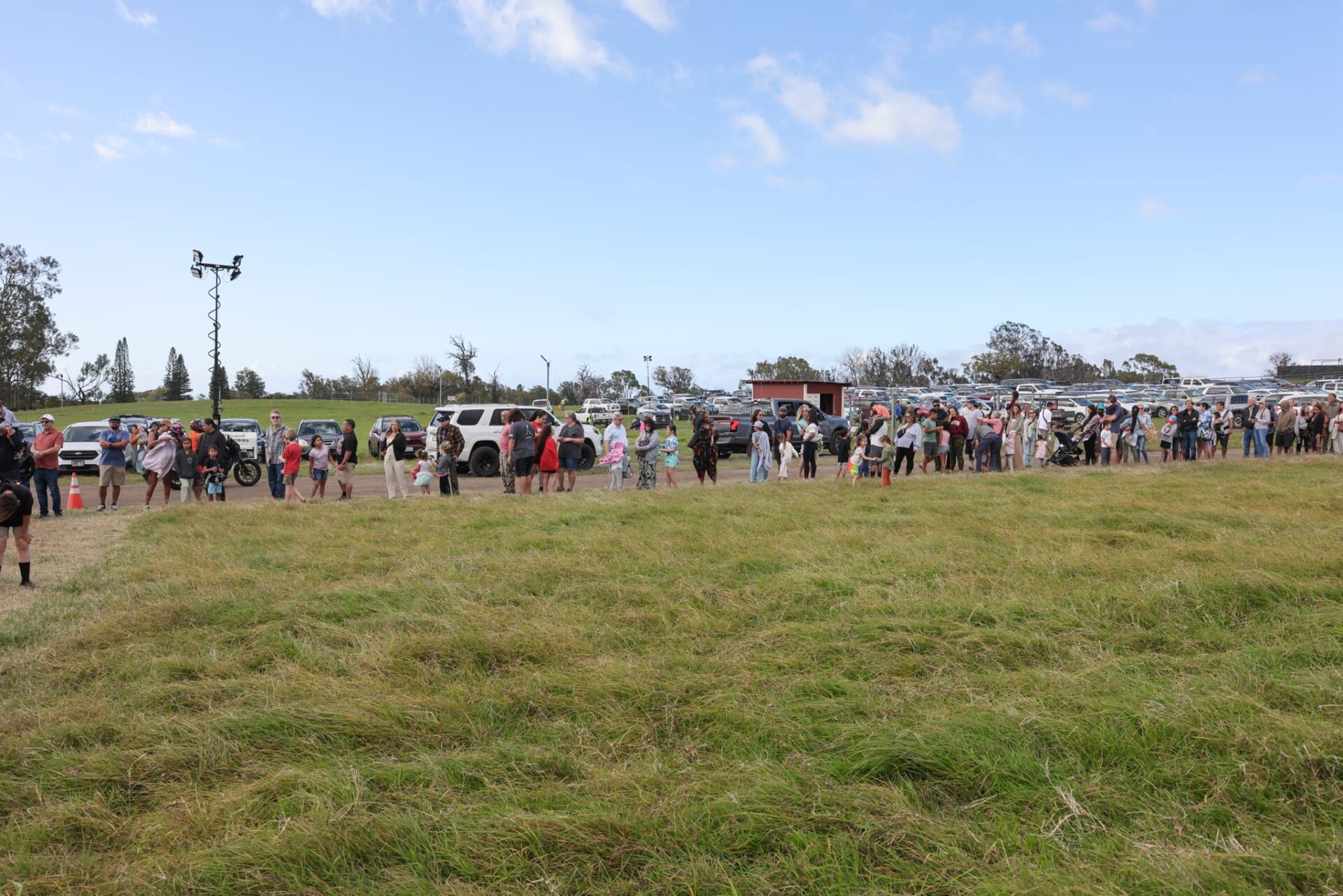 A long line of people stands in a grassy field at the Oskie Rice Event Center, with cars and trees in the background under a blue sky, adding to the vibrant atmosphere of Maui events.